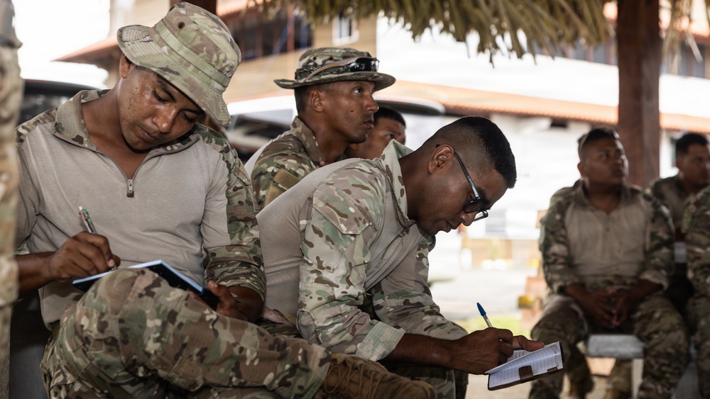 A group of military personnel in camouflage uniforms are gathered outdoors in what appears to be a training or briefing session. Several soldiers in the foreground are actively taking notes on small notepads or booklets. The setting appears to be a tropical or subtropical location based on the palm-thatched structure and vegetation visible in the background.