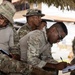 A group of military personnel in camouflage uniforms are gathered outdoors in what appears to be a training or briefing session. Several soldiers in the foreground are actively taking notes on small notepads or booklets. The setting appears to be a tropical or subtropical location based on the palm-thatched structure and vegetation visible in the background.