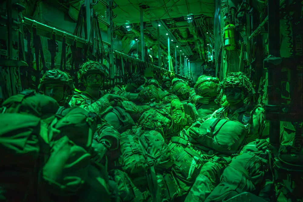 A group of heavily equipped military personnel are seated inside the cargo hold of a military transport aircraft, illuminated by green lighting. The soldiers are wearing full combat gear including helmets, body armor, and large rucksacks, suggesting preparation for an airborne or air assault operation. The green lighting is consistent with night operations lighting used in military aircraft.