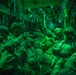 A group of heavily equipped military personnel are seated inside the cargo hold of a military transport aircraft, illuminated by green lighting. The soldiers are wearing full combat gear including helmets, body armor, and large rucksacks, suggesting preparation for an airborne or air assault operation. The green lighting is consistent with night operations lighting used in military aircraft.