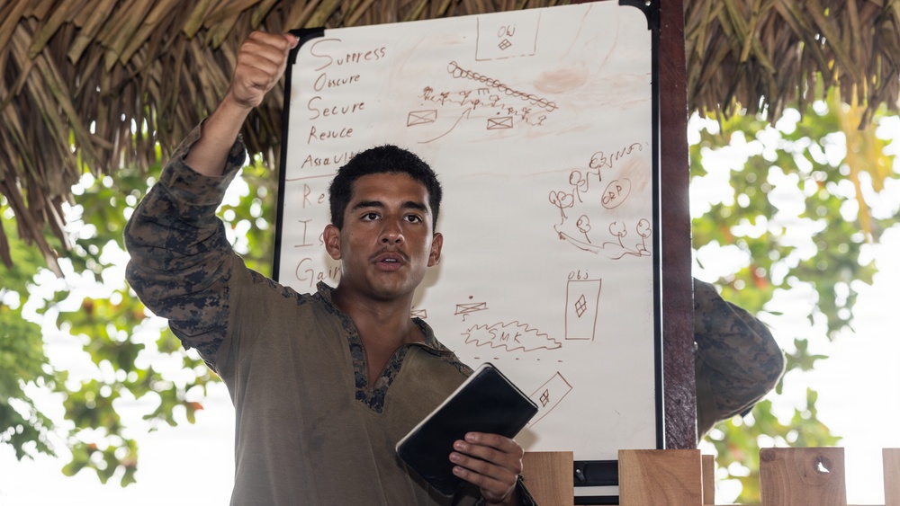 A person in military camouflage uniform is presenting a briefing under a thatched-roof structure in what appears to be a tropical outdoor setting. They are holding a tablet device and gesturing toward a whiteboard that contains written text and tactical diagrams. The setting and attire suggest a military field training or operational planning session.
