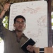 A person in military camouflage uniform is presenting a briefing under a thatched-roof structure in what appears to be a tropical outdoor setting. They are holding a tablet device and gesturing toward a whiteboard that contains written text and tactical diagrams. The setting and attire suggest a military field training or operational planning session.