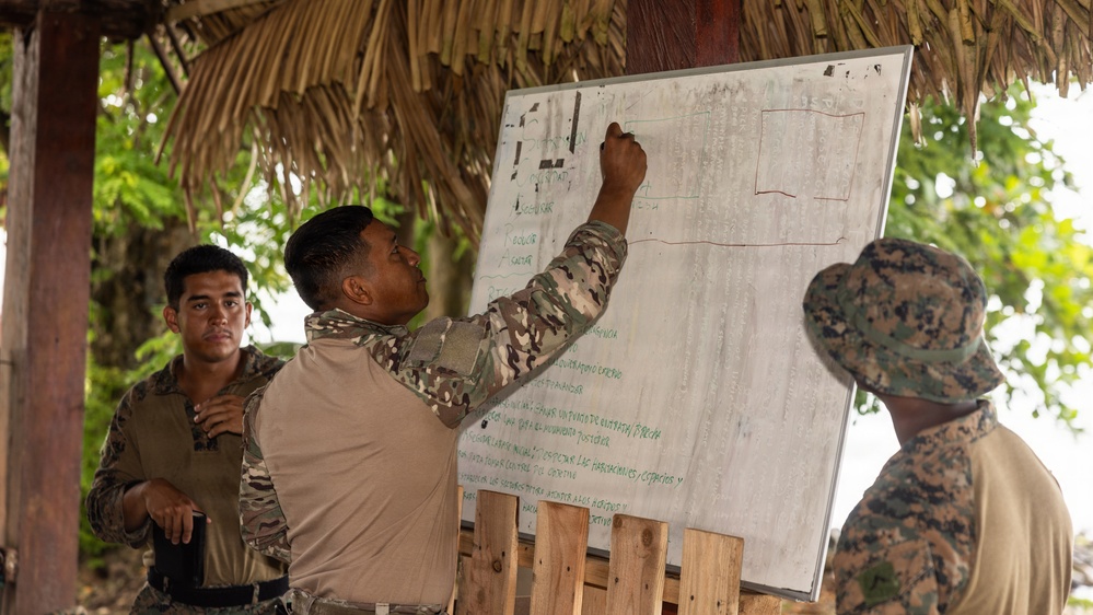 Three military personnel are gathered under a thatched-roof open-air structure for what appears to be a briefing or planning session. One soldier in camouflage uniform is writing or pointing at a whiteboard that contains text in Spanish and a drawn diagram. The setting suggests a field environment, possibly in a tropical or Latin American location.