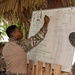 Three military personnel are gathered under a thatched-roof open-air structure for what appears to be a briefing or planning session. One soldier in camouflage uniform is writing or pointing at a whiteboard that contains text in Spanish and a drawn diagram. The setting suggests a field environment, possibly in a tropical or Latin American location.