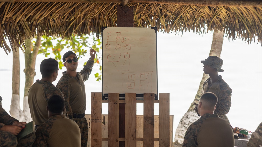 Military personnel in camouflage uniforms are gathered under a thatched-roof open-air structure near a beach or coastal area. One individual is presenting at a whiteboard mounted on a wooden pallet stand, pointing to diagrams that appear to show organizational or tactical charts. The setting suggests a field briefing or planning session in a tropical environment.