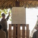 Military personnel in camouflage uniforms are gathered under a thatched-roof open-air structure near a beach or coastal area. One individual is presenting at a whiteboard mounted on a wooden pallet stand, pointing to diagrams that appear to show organizational or tactical charts. The setting suggests a field briefing or planning session in a tropical environment.