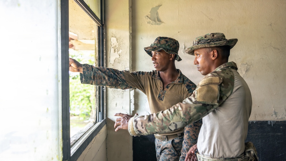 Two military personnel in different camouflage uniforms are standing inside a weathered building, looking out through a window. The individual on the left, wearing MARPAT (Marine pattern) camouflage and a patrol cap, is pointing toward something outside. The scene appears to be a training or reconnaissance exercise in a tropical or humid environment.