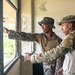 Two military personnel in different camouflage uniforms are standing inside a weathered building, looking out through a window. The individual on the left, wearing MARPAT (Marine pattern) camouflage and a patrol cap, is pointing toward something outside. The scene appears to be a training or reconnaissance exercise in a tropical or humid environment.