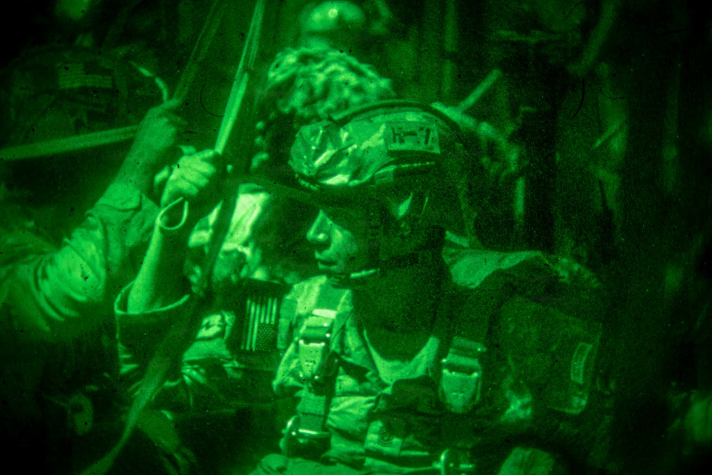 A night-vision captured image showing a military personnel in full tactical gear inside what appears to be a military aircraft or vehicle. The soldier is wearing a combat helmet, body armor, and has a visible US flag patch on their uniform. The characteristic green monochrome coloring is consistent with night-vision goggle (NVG) photography.