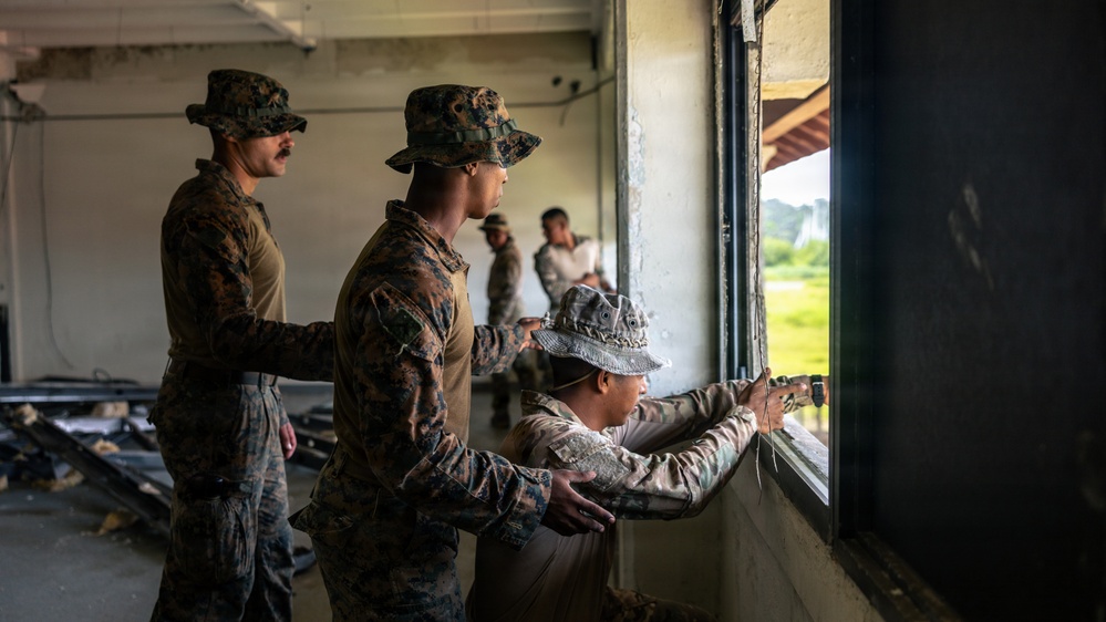 Military personnel in camouflage uniforms and boonie hats are working inside a partially demolished or under-renovation building. They appear to be conducting construction or repair work near a large window opening, with some personnel working on the window frame. The setting appears to be a tropical or subtropical location based on the vegetation visible outside.