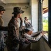 Military personnel in camouflage uniforms and boonie hats are working inside a partially demolished or under-renovation building. They appear to be conducting construction or repair work near a large window opening, with some personnel working on the window frame. The setting appears to be a tropical or subtropical location based on the vegetation visible outside.