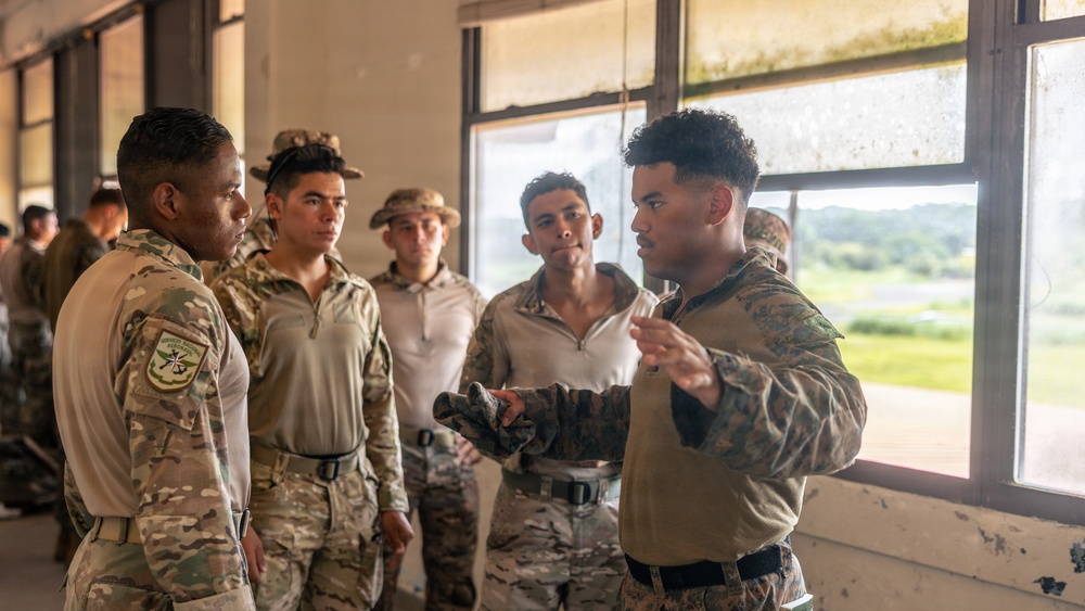 A group of military personnel in camouflage uniforms are gathered inside what appears to be a weathered building, engaged in a briefing or instructional discussion. One individual on the right, wearing a distinct camouflage pattern consistent with U.S. Marine Corps MARPAT, appears to be addressing or instructing the others. The setting and mixed uniform styles suggest a multinational training exercise or joint military operation.