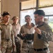 A group of military personnel in camouflage uniforms are gathered inside what appears to be a weathered building, engaged in a briefing or instructional discussion. One individual on the right, wearing a distinct camouflage pattern consistent with U.S. Marine Corps MARPAT, appears to be addressing or instructing the others. The setting and mixed uniform styles suggest a multinational training exercise or joint military operation.