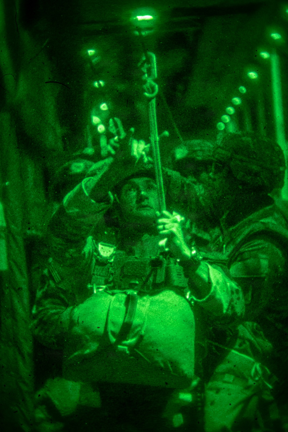 A night-vision captured image showing military personnel inside what appears to be a military aircraft, likely preparing for or conducting an airborne operation. The green monochromatic imagery is characteristic of night-vision device (NVD) photography. Personnel are wearing tactical gear and appear to be attaching or checking static lines or jump equipment overhead.