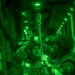 A night-vision captured image showing military personnel inside what appears to be a military aircraft, likely preparing for or conducting an airborne operation. The green monochromatic imagery is characteristic of night-vision device (NVD) photography. Personnel are wearing tactical gear and appear to be attaching or checking static lines or jump equipment overhead.
