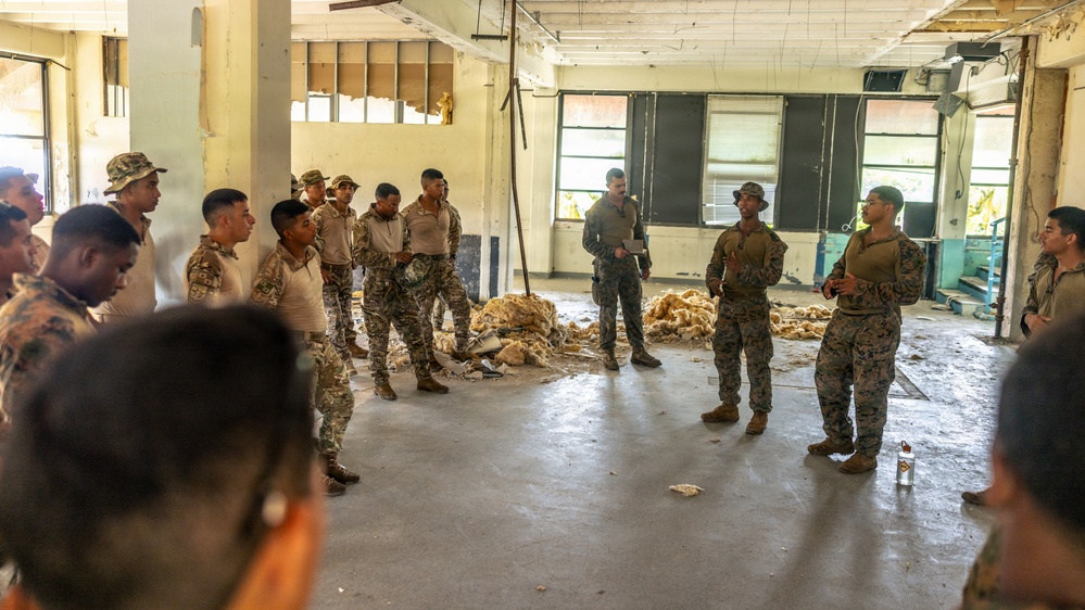 A group of military personnel in camouflage uniforms are gathered inside a dilapidated, abandoned building for what appears to be a briefing or instruction session. Two individuals in the center appear to be addressing the larger group, with one holding a document or clipboard. The mixed uniform patterns suggest personnel from potentially two different military forces participating in a joint exercise or training event.