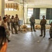 A group of military personnel in camouflage uniforms are gathered inside a dilapidated, abandoned building for what appears to be a briefing or instruction session. Two individuals in the center appear to be addressing the larger group, with one holding a document or clipboard. The mixed uniform patterns suggest personnel from potentially two different military forces participating in a joint exercise or training event.