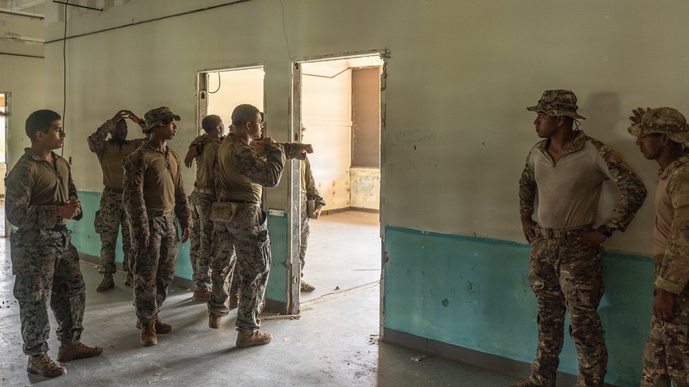 A group of military personnel in mixed camouflage uniforms are gathered inside a worn, partially deteriorated building. One individual appears to be instructing or briefing the group near a doorway, pointing toward an adjacent room. The setting suggests a tactical training exercise or building clearance drill.