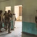 A group of military personnel in mixed camouflage uniforms are gathered inside a worn, partially deteriorated building. One individual appears to be instructing or briefing the group near a doorway, pointing toward an adjacent room. The setting suggests a tactical training exercise or building clearance drill.
