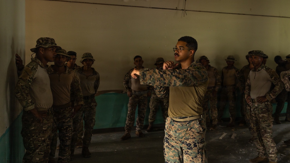 A military instructor in MARPAT (US Marine Corps digital camouflage) uniform is demonstrating a technique, likely a combat or martial arts move, to a group of soldiers wearing a different camouflage pattern (multicam-style). The setting appears to be an indoor training facility or building with bare concrete walls. The instructor appears to be leading a close-combat or hand-to-hand training session.