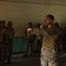A military instructor in MARPAT (US Marine Corps digital camouflage) uniform is demonstrating a technique, likely a combat or martial arts move, to a group of soldiers wearing a different camouflage pattern (multicam-style). The setting appears to be an indoor training facility or building with bare concrete walls. The instructor appears to be leading a close-combat or hand-to-hand training session.
