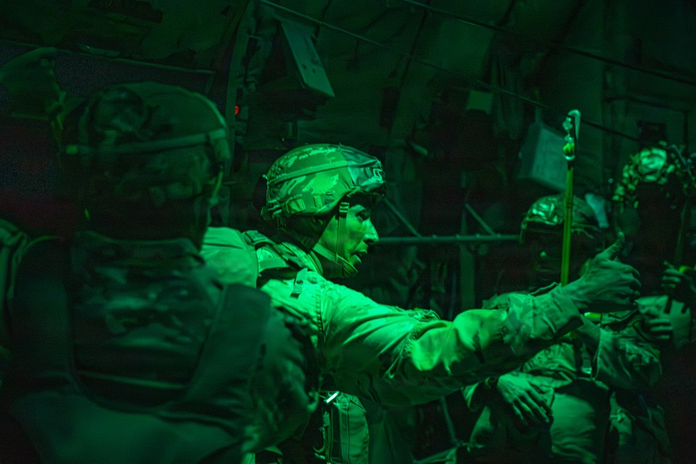 Military personnel are seated inside what appears to be a military transport aircraft, illuminated by green cabin lighting typical of night operations. The soldiers are in full combat gear including tactical helmets and body armor, with one central figure reaching toward what appears to be a static line or anchor cable overhead. The scene is consistent with pre-jump preparation for a military airborne/parachute operation.