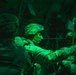 Military personnel are seated inside what appears to be a military transport aircraft, illuminated by green cabin lighting typical of night operations. The soldiers are in full combat gear including tactical helmets and body armor, with one central figure reaching toward what appears to be a static line or anchor cable overhead. The scene is consistent with pre-jump preparation for a military airborne/parachute operation.