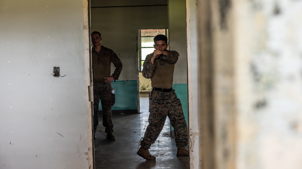 Two military personnel in MARPAT (Marine pattern) camouflage uniforms are inside a dilapidated building, appearing to conduct close-quarters combat or martial arts training. The foreground figure is in a fighting stance with fists raised, while the second individual stands observing in the background. The setting appears to be an abandoned or training facility structure.