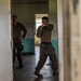 Two military personnel in MARPAT (Marine pattern) camouflage uniforms are inside a dilapidated building, appearing to conduct close-quarters combat or martial arts training. The foreground figure is in a fighting stance with fists raised, while the second individual stands observing in the background. The setting appears to be an abandoned or training facility structure.