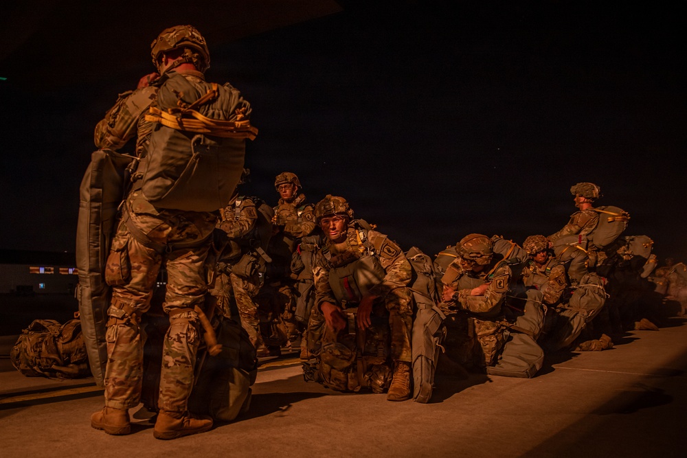 A group of military personnel in full combat gear and camouflage uniforms are staged on what appears to be a tarmac or airfield at night. The soldiers are equipped with large rucksacks, helmets, and parachute equipment, suggesting preparation for an airborne operation. The scene is dramatically lit with warm orange ambient light against a dark night sky.