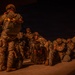 A group of military personnel in full combat gear and camouflage uniforms are staged on what appears to be a tarmac or airfield at night. The soldiers are equipped with large rucksacks, helmets, and parachute equipment, suggesting preparation for an airborne operation. The scene is dramatically lit with warm orange ambient light against a dark night sky.