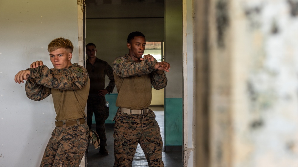 Two military personnel in MARPAT (Marine pattern) camouflage uniforms are practicing combat or martial arts techniques in a doorway of a building. They appear to be performing synchronized defensive or striking stances with arms raised and crossed. A third individual in darker uniform is visible in the background observing.