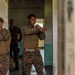 Two military personnel in MARPAT (Marine pattern) camouflage uniforms are practicing combat or martial arts techniques in a doorway of a building. They appear to be performing synchronized defensive or striking stances with arms raised and crossed. A third individual in darker uniform is visible in the background observing.
