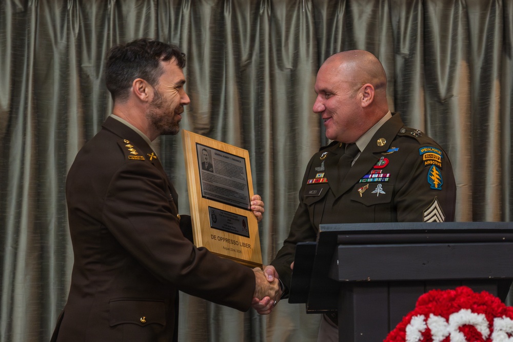 Two military personnel in U.S. Army dress uniforms are shaking hands during a formal ceremony. One soldier is presenting a wooden commemorative plaque to the other, which bears the motto 'DE OPPRESSO LIBER' — the motto of the U.S. Army Special Forces. The recipient on the right wears Special Forces, Ranger, and Airborne tabs along with multiple service ribbons and badges.