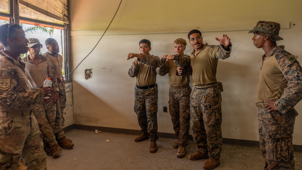 A group of military personnel in camouflage uniforms are gathered inside a worn interior room, appearing to conduct a close-quarters combat or martial arts training session. Two individuals in the center are demonstrating fighting stances or techniques while others observe. The group includes personnel in both MARPAT (Marine pattern) and tan/khaki uniforms, suggesting a joint or mixed-unit training exercise.