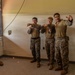 A group of military personnel in camouflage uniforms are gathered inside a worn interior room, appearing to conduct a close-quarters combat or martial arts training session. Two individuals in the center are demonstrating fighting stances or techniques while others observe. The group includes personnel in both MARPAT (Marine pattern) and tan/khaki uniforms, suggesting a joint or mixed-unit training exercise.