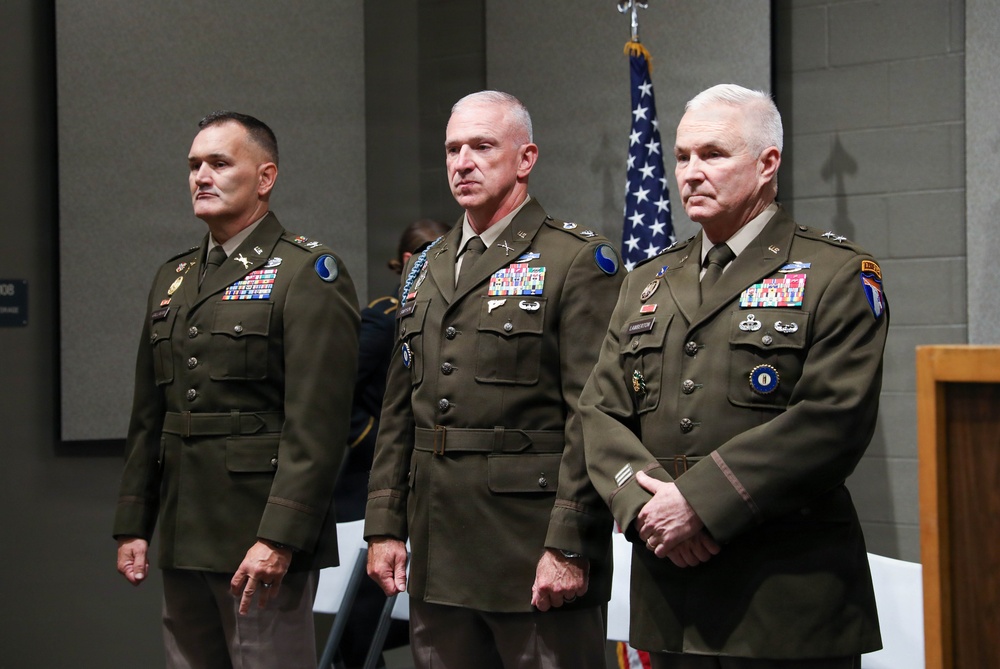 Three senior U.S. Army officers in dress green uniforms stand at attention during what appears to be a formal military ceremony. All three wear extensive medal and ribbon arrays indicating long service careers, with rank insignia visible on their shoulders. An American flag is displayed in the background.