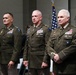 Three senior U.S. Army officers in dress green uniforms stand at attention during what appears to be a formal military ceremony. All three wear extensive medal and ribbon arrays indicating long service careers, with rank insignia visible on their shoulders. An American flag is displayed in the background.