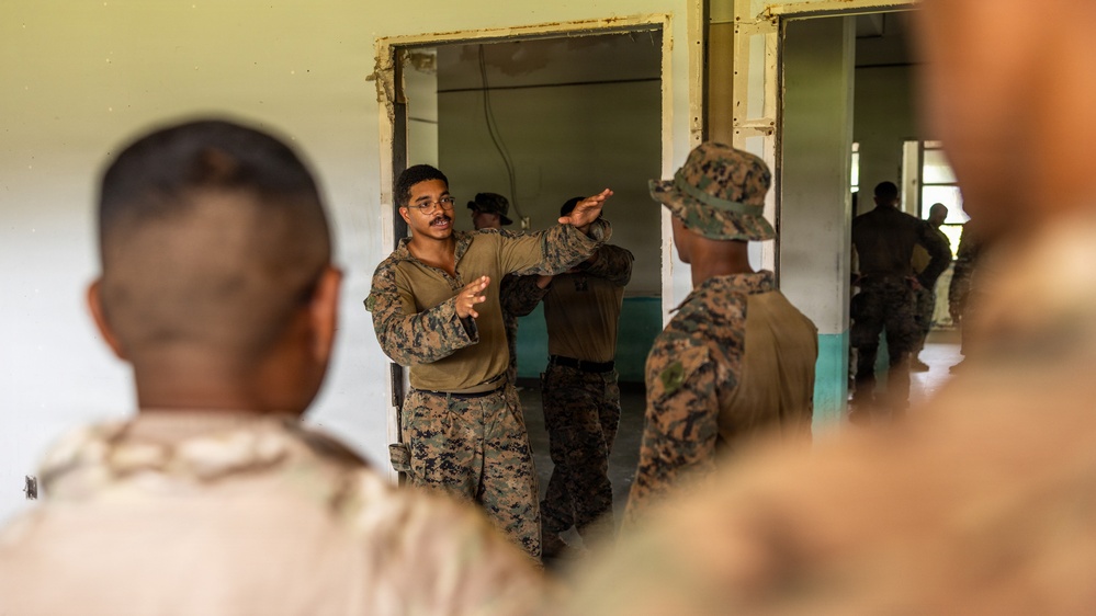 Military personnel in camouflage uniforms appear to be engaged in a training or briefing session inside a building. A central figure wearing glasses and MARPAT digital camouflage is gesturing with both hands, apparently instructing or demonstrating something to others. The setting appears to be a worn or partially renovated building used as a training facility.
