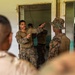 Military personnel in camouflage uniforms appear to be engaged in a training or briefing session inside a building. A central figure wearing glasses and MARPAT digital camouflage is gesturing with both hands, apparently instructing or demonstrating something to others. The setting appears to be a worn or partially renovated building used as a training facility.