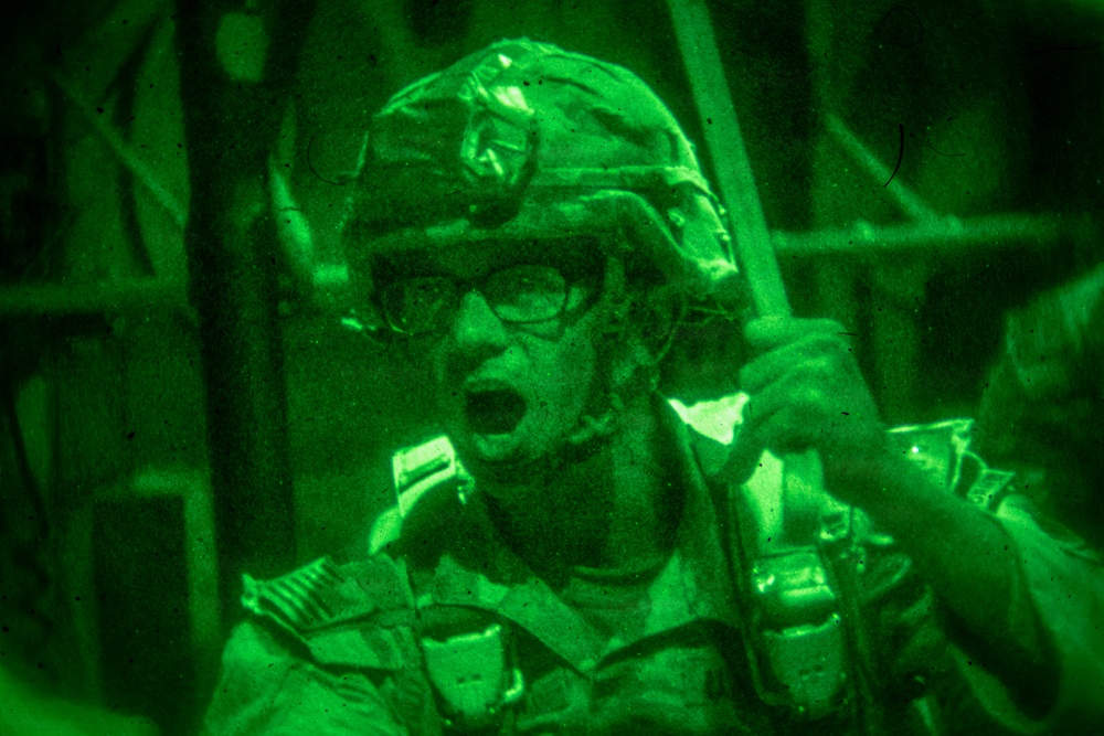 A night-vision captured image showing a military personnel wearing a combat helmet and glasses, appearing to be shouting or giving commands. The image is rendered entirely in green monochrome tones characteristic of night-vision optics. The soldier appears to be holding a piece of equipment, possibly a radio handset or weapon component.