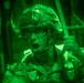 A night-vision captured image showing a military personnel wearing a combat helmet and glasses, appearing to be shouting or giving commands. The image is rendered entirely in green monochrome tones characteristic of night-vision optics. The soldier appears to be holding a piece of equipment, possibly a radio handset or weapon component.