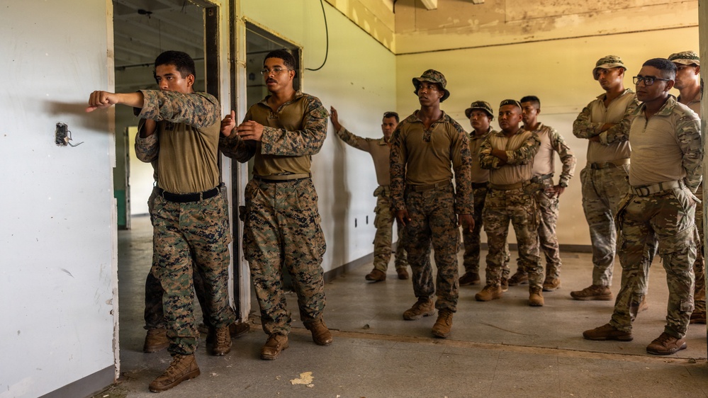 A group of military personnel in various camouflage uniforms are participating in what appears to be a close-quarters battle (CQB) or room-clearing training exercise inside a building. Two soldiers at the front are demonstrating a door-entry technique while a larger group observes from behind. The mixed uniform patterns suggest this may be a joint or multinational training exercise.