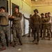 A group of military personnel in various camouflage uniforms are participating in what appears to be a close-quarters battle (CQB) or room-clearing training exercise inside a building. Two soldiers at the front are demonstrating a door-entry technique while a larger group observes from behind. The mixed uniform patterns suggest this may be a joint or multinational training exercise.