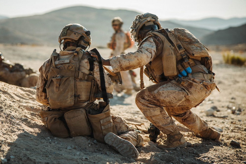 Two soldiers in desert camouflage uniforms are engaged in what appears to be a tactical combat casualty care (TCCC) or medical training exercise in an arid, desert environment. One soldier is kneeling on the ground while a second soldier, wearing blue medical gloves, attends to their back or equipment. A third individual in civilian-style clothing is visible in the blurred background.
