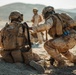 Two soldiers in desert camouflage uniforms are engaged in what appears to be a tactical combat casualty care (TCCC) or medical training exercise in an arid, desert environment. One soldier is kneeling on the ground while a second soldier, wearing blue medical gloves, attends to their back or equipment. A third individual in civilian-style clothing is visible in the blurred background.