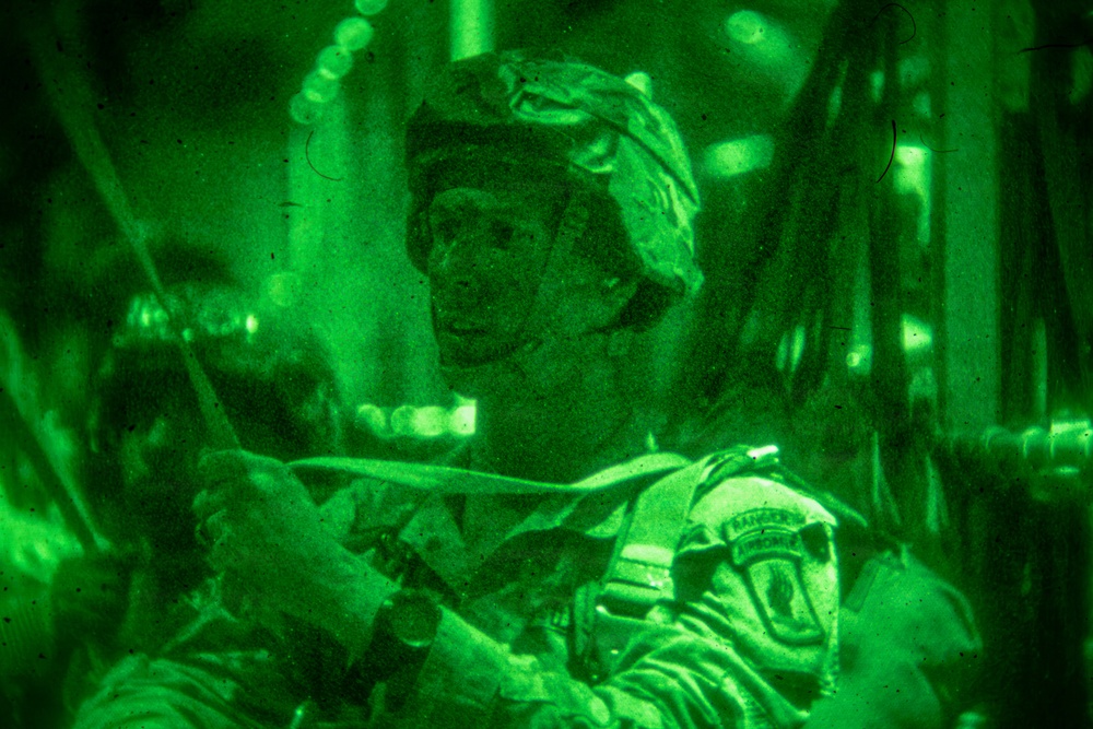 A night-vision photograph showing a military soldier wearing combat gear and a patrol cap inside what appears to be a military aircraft. The image is captured entirely in the characteristic green monochrome of night-vision optics. A shoulder patch is visible on the soldier's uniform, appearing to read 'Ranger' and 'Airborne'.