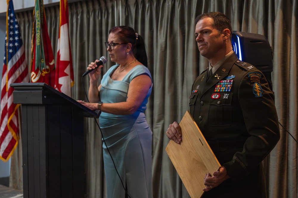A military ceremony featuring a U.S. Army officer in dress uniform standing beside a civilian woman who is speaking at a podium with a microphone. The officer holds a wooden plaque or award and wears an extensively decorated uniform with numerous ribbons, badges, and Special Forces/Airborne patches. Multiple national and military flags are visible in the background, including the U.S. and Canadian flags.