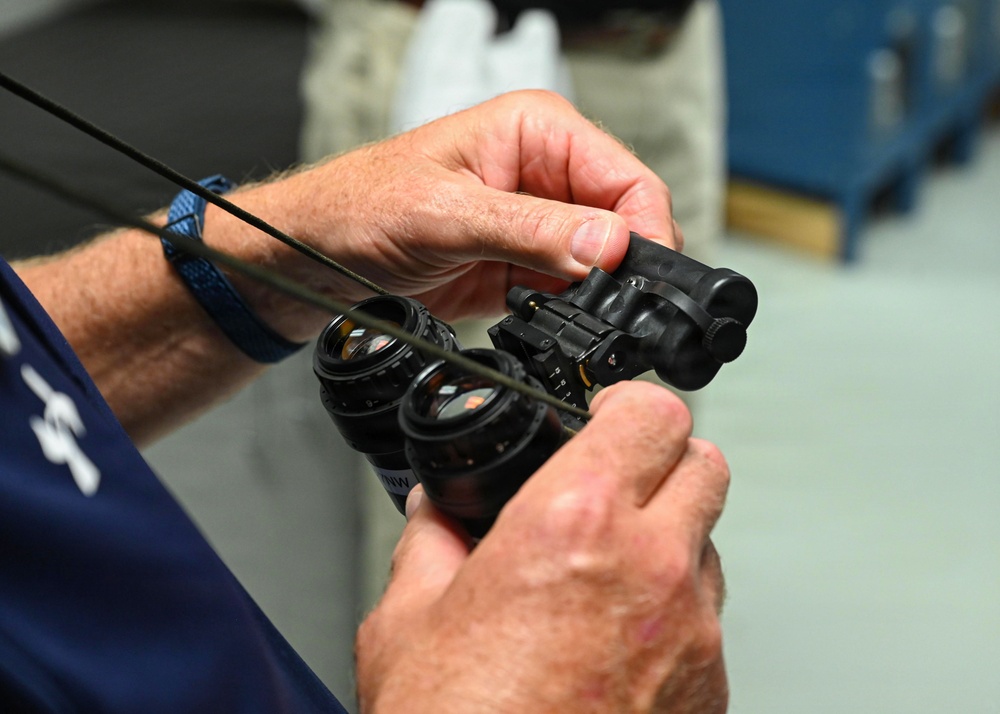 A person is handling a pair of night vision goggles (NVGs) in what appears to be a maintenance or inspection facility. The individual is manipulating the dual-tube optical device with both hands, possibly performing assembly, disassembly, or inspection work. The setting appears to be a technical workshop or equipment maintenance area.
