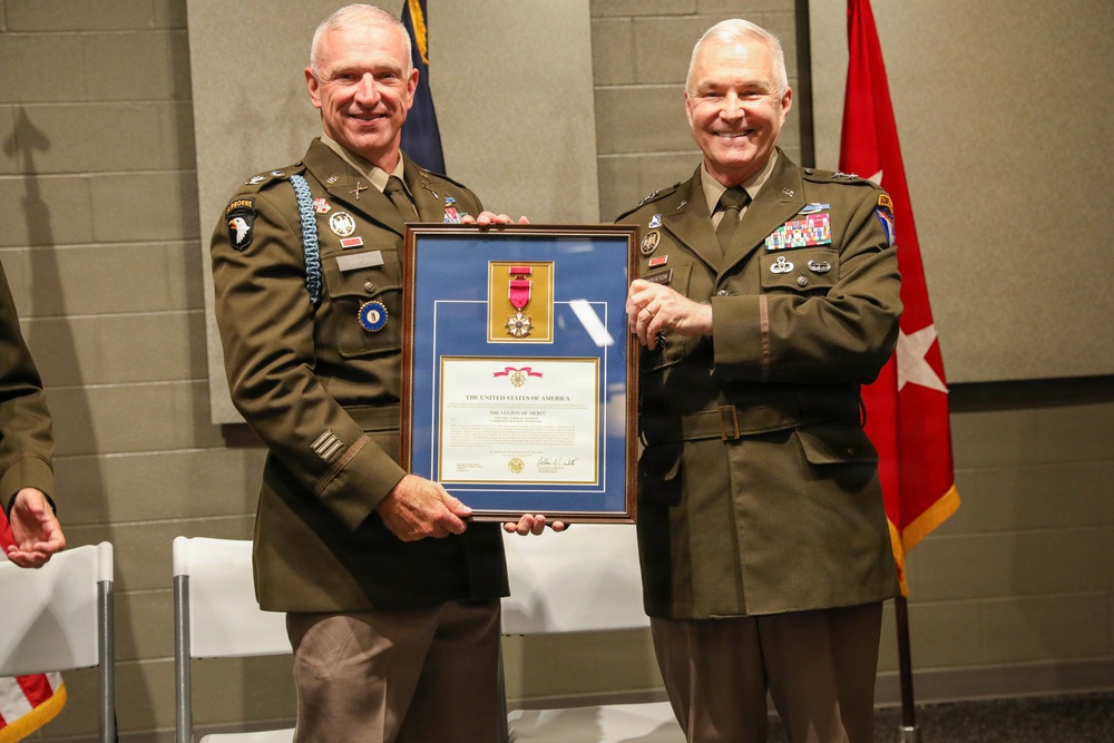 Two U.S. Army officers in service uniforms are posing together while holding a framed award certificate. The framed display contains a medal with a red ribbon and an official certificate reading 'The United States of America' and 'The Legion of Merit.' The ceremony appears to be a formal military awards presentation.